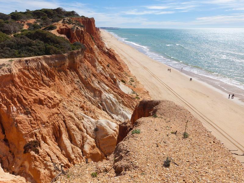 Scogliere rosse che dominano una larga spiaggia sabbiosa con pochi pedoni.