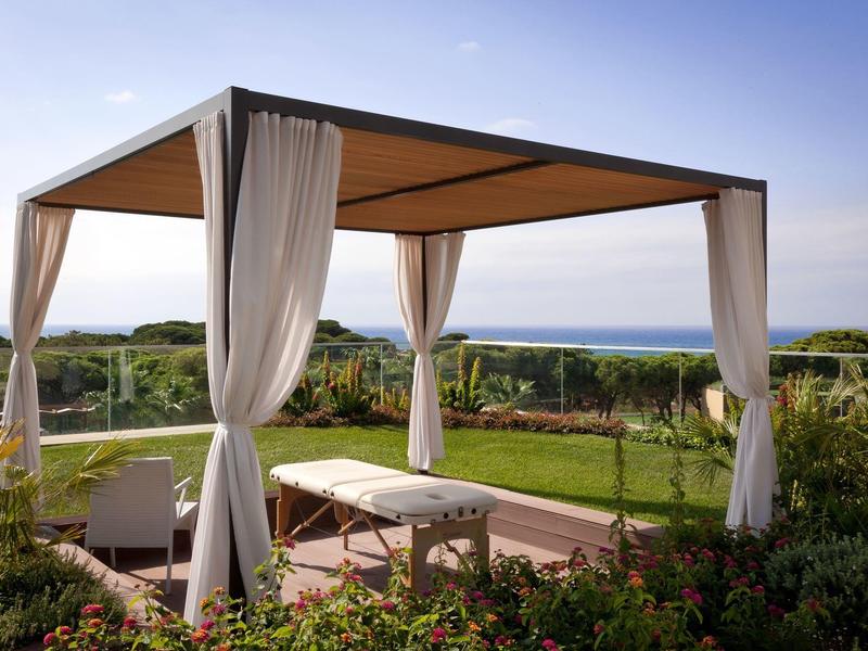 Padiglione moderno con tende bianche in un'oasi di giardino con vista mare sotto un cielo sereno.