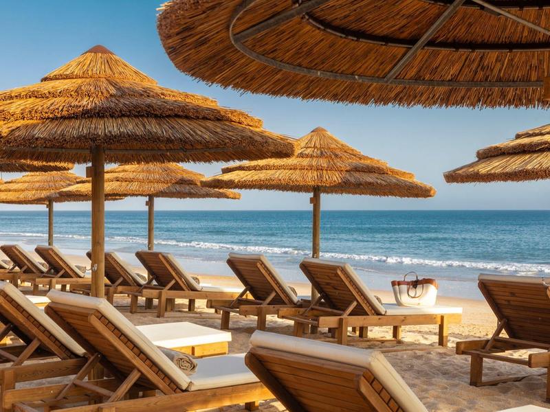 File di lettini in legno sotto ombrelloni di paglia su una spiaggia sabbiosa con vista sul mare.