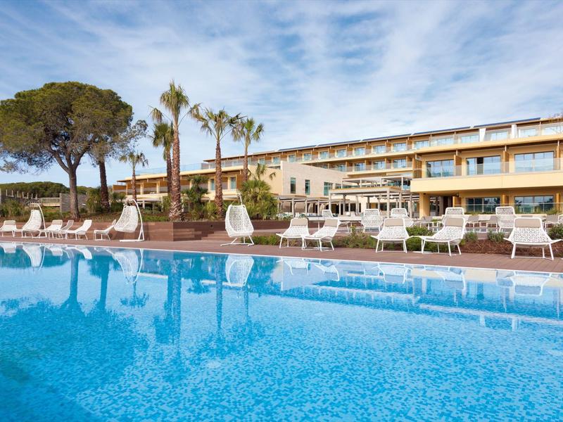 Grande piscina con lettini davanti a un hotel su più piani e palme sotto un cielo blu.