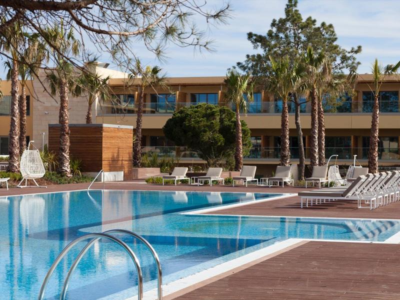 Piscina esterna moderna con lettini e alberi davanti a un edificio dell'hotel.