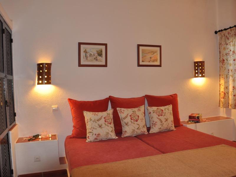 Gemütliches Schlafzimmer mit rotem Bett, drei Kissen, floralem Vorhang und zwei Wandlampen.