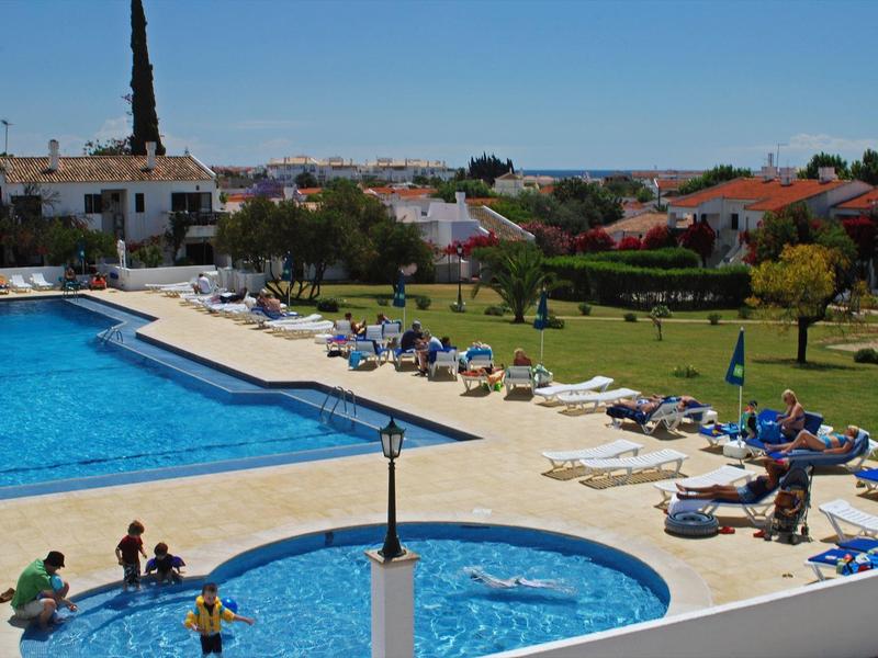 Großer Außenpool mit Sonnenliegen und Menschen, umgeben von Häusern und grünem Garten bei klarem Himmel.