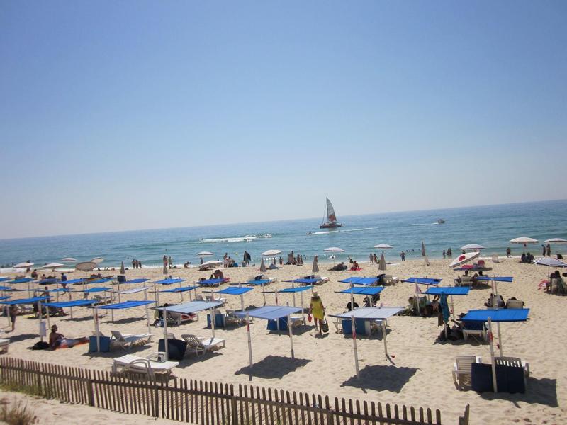 Strand mit vielen blauen Liegestühlen und Sonnenschirmen, ruhiges Meer und Segelboot im Hintergrund.