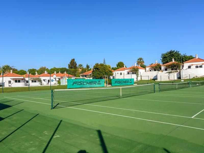 Leerer grüner Tennisplatz mit Netz vor weißen Häusern unter klarem blauem Himmel.