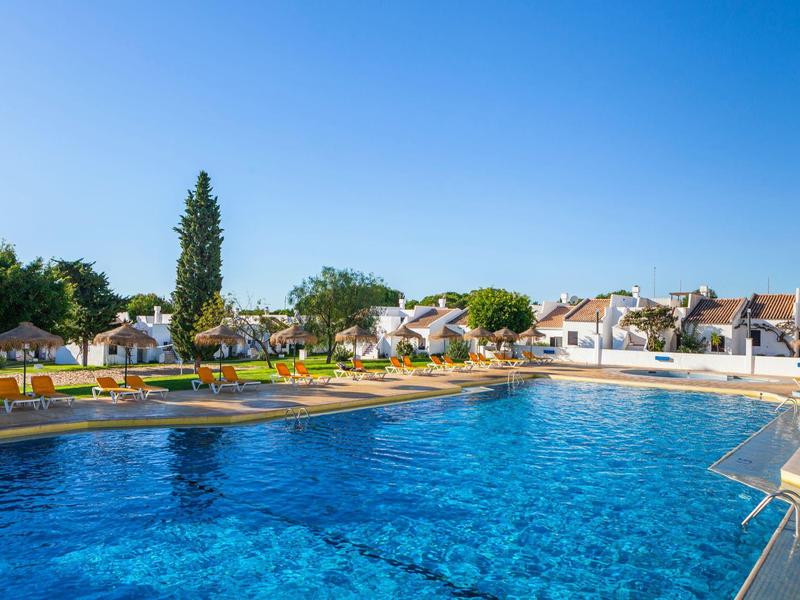 Großer blauer Pool mit Liegestühlen und weißen Häusern im Hintergrund bei klarem Himmel.