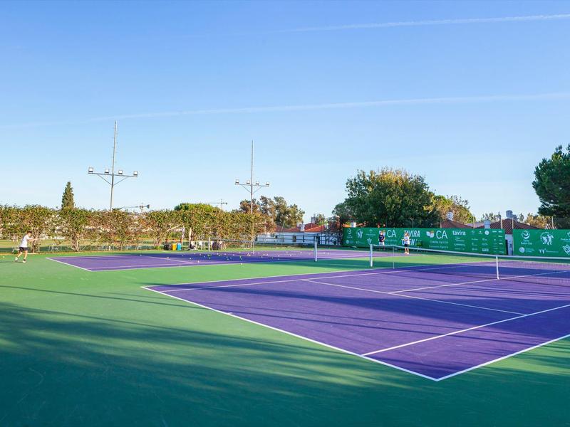 Tennisplatz mit grünem und violettem Belag, umgeben von Bäumen und strahlend blauem Himmel.