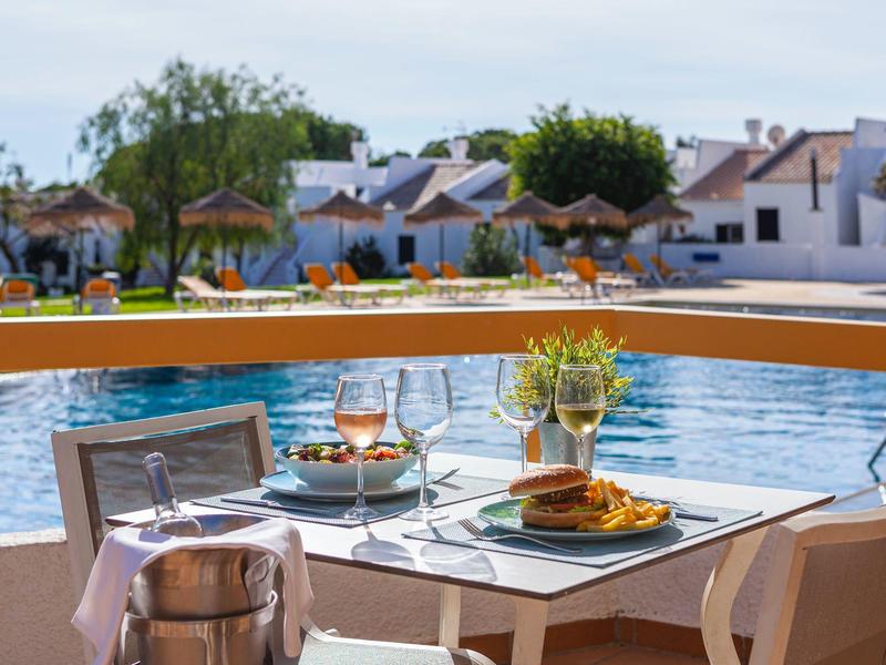 Tisch mit Frühstück neben hellem Pool, umgeben von Häusern und Bäumen im Hintergrund.