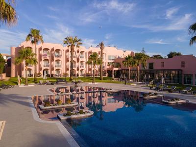 Hotelgebäude mit rosa Fassade, umgeben von Palmen und einem großen Pool mit Liegestühlen.