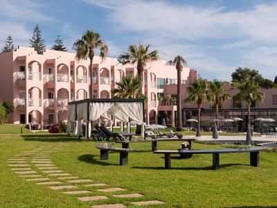 Hotelgebäude mit rosa Fassade, Palmen und grüner Rasenfläche mit Sitzbänken und Wegen.