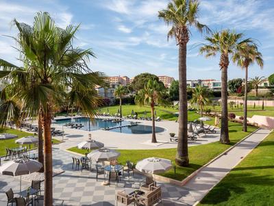 Poolbereich mit Sonnenschirmen, Palmen und gepflegtem Rasen in einer Hotelanlage.