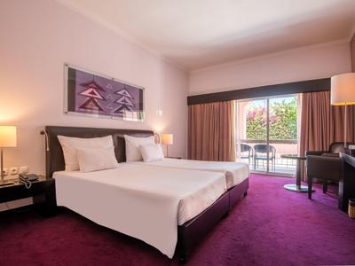 Modernes Hotelzimmer mit großem Bett, Teppichboden und Balkon mit Blick auf grüne Bäume.