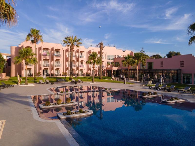 Hotelgebäude mit rosa Fassade, umgeben von Palmen und einem großen Pool mit Liegestühlen.