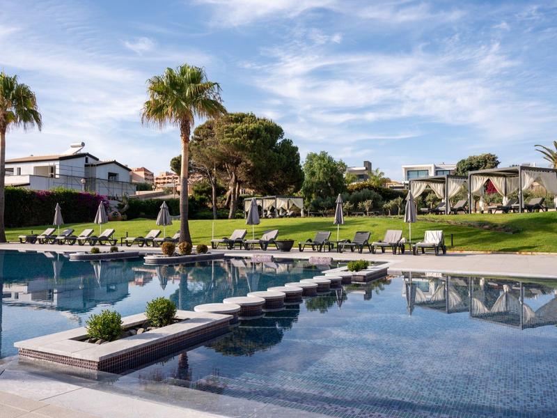 Moderner Hotelpool mit Palmen und Liegestühlen unter blauem Himmel.