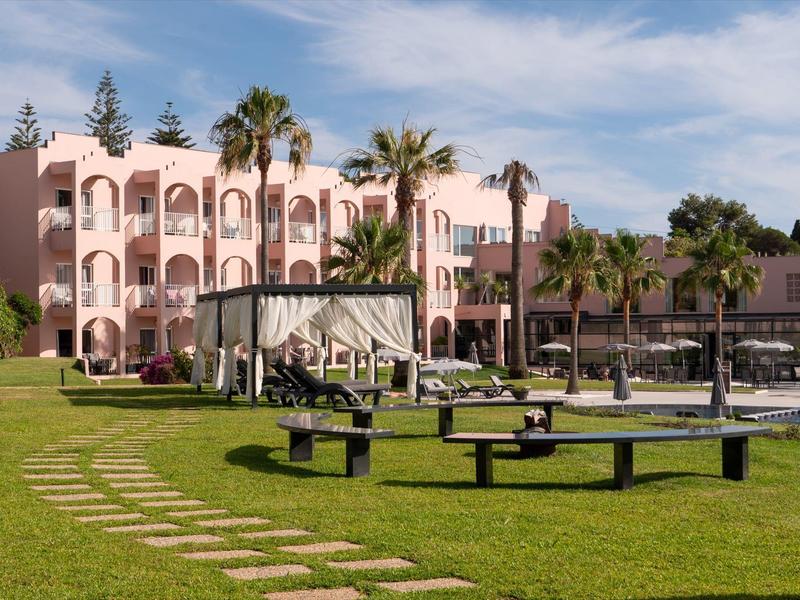 Hotelgebäude mit rosa Fassade, Palmen und grüner Rasenfläche mit Sitzbänken und Wegen.