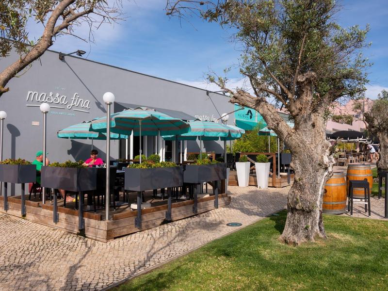 Außenbereich eines Cafés mit grünen Sonnenschirmen und Tischen neben einem Baum und Rasenfläche.