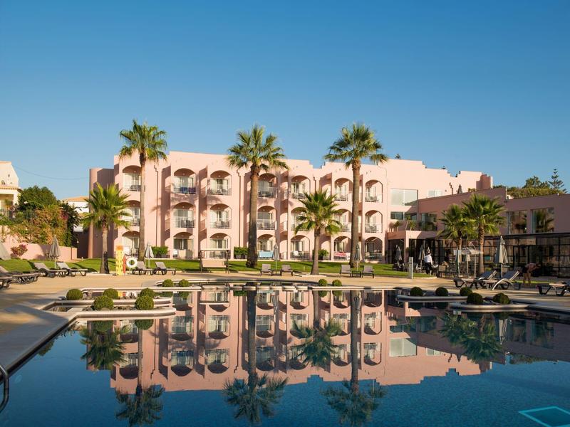 Hotelgebäude mit Balkonen und Palmen spiegelt sich in klarem Poolwasser unter blauem Himmel.