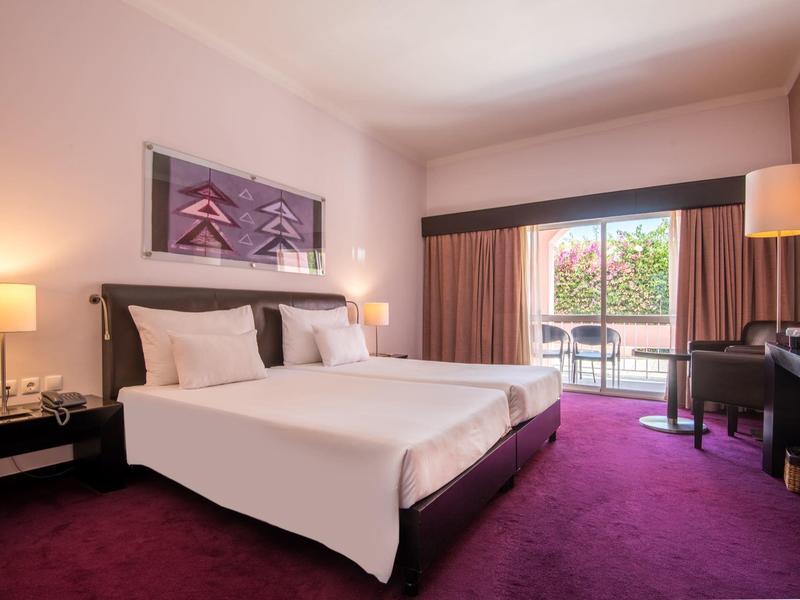 Modernes Hotelzimmer mit großem Bett, Teppichboden und Balkon mit Blick auf grüne Bäume.