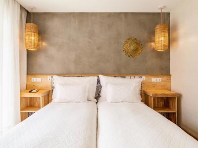 Zwei Einzelbetten mit weißen Bettlaken, Holznachtische, graue Wand und hängende Lampen aus Korbgeflecht.