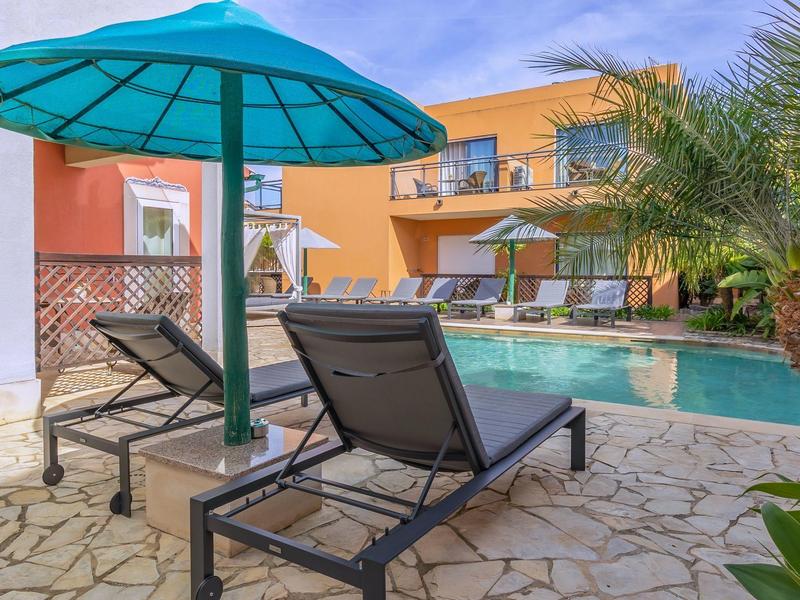 Poolbereich mit Sonnenliegen, Sonnenschirmen und bunten zwei-geschossigen Gebäuden im Hintergrund.