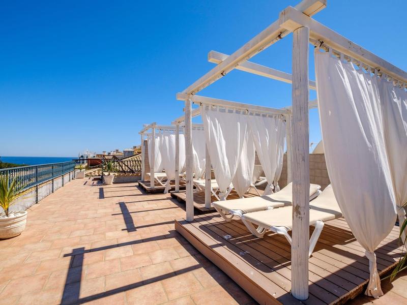 Terrassenbereich mit weißen Baldachin-Liegen, blauer Himmel und Meer am Horizont.