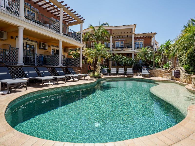 Schwimmbad mit klarem Wasser, umgeben von Liegestühlen und mediterranen Gebäuden unter blauem Himmel.