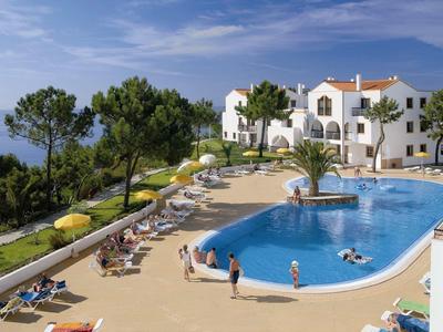 Ein blauer Pool mit Sonnenschirmen, Liegestühlen und weißen Gebäuden im Hintergrund.