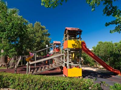Bunter Spielplatz mit Rutsche, Klettergerüst und blühenden Büschen unter blauem Himmel.