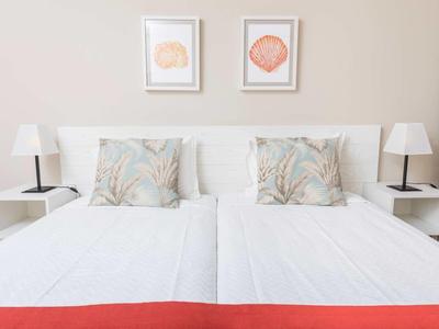 Hell eingerichtetes Schlafzimmer mit weißem Doppelbett, zwei Lampen und maritimen Bildern an der Wand.