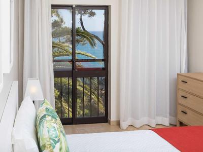 Helles Schlafzimmer mit weißen Wänden, Bett, großer Fensterfront und Blick auf Palmen und Meer.