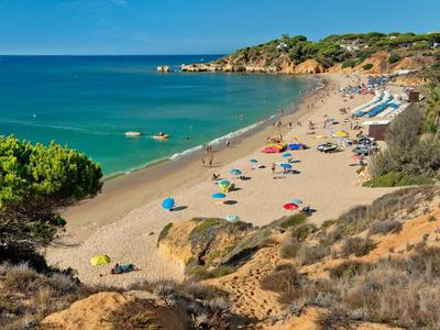 Sandstrand mit bunten Sonnenschirmen, blauem Meer und bewachsenen Klippen unter klarem Himmel.