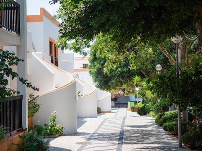 Gepflasterter Weg umgeben von weißen Häusern und grünen Bäumen bei Sonnenschein.