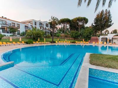 Großer blauer Pool mit gelben Liegestühlen, umgeben von grünen Bäumen und weißen Gebäuden.