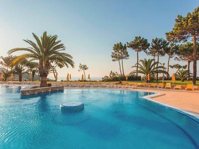 Großer blauer Pool mit Palmen davor und klarem Himmel im Hintergrund, sonnige Atmosphäre.
