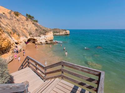 Holzsteg führt zu kleinem Sandstrand mit Menschen, Felsen und klarem blauem Meer unter blauem Himmel.