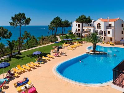 Großer blauer Pool mit gelben Liegestühlen und weißen Gebäuden am Meer unter blauem Himmel.