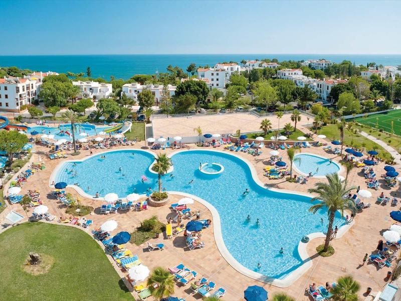 Großer Hotelpool mit Sonnenschirmen, Liegen und Palmen nahe dem Meer und umgeben von Gebäuden.