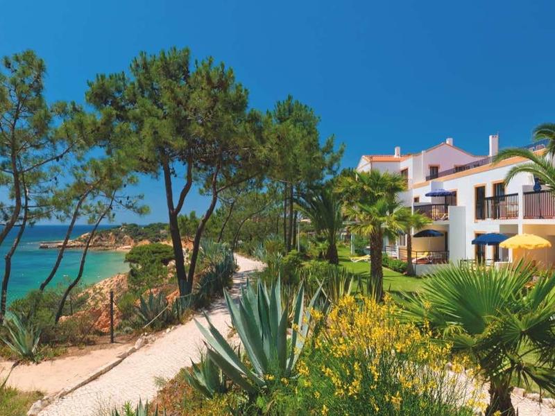 Grünes Gartenhotel mit Palmen, Sandweg und Blick auf das blaue Meer unter klarem Himmel.