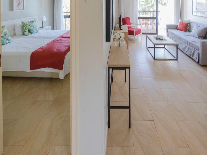 Heller Wohnbereich mit grauem Sofa, roten Akzenten und angrenzendem Schlafzimmer mit roter Bettdecke.