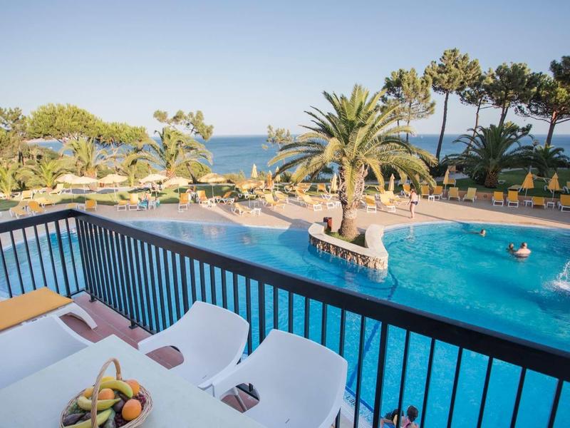 Blick auf einen blauen Pool mit Palmen, Stühlen und Meer im Hintergrund an sonnigem Tag.