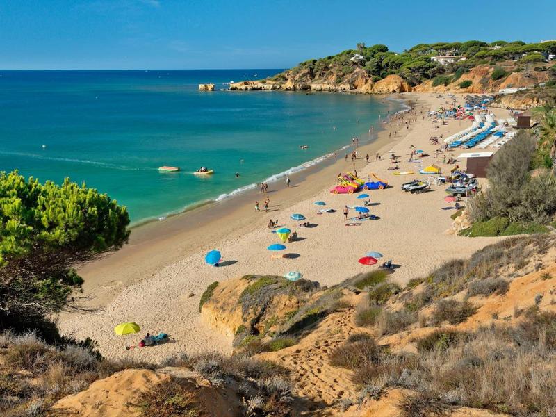 Sandstrand mit bunten Sonnenschirmen, blauem Meer und bewachsenen Klippen unter klarem Himmel.