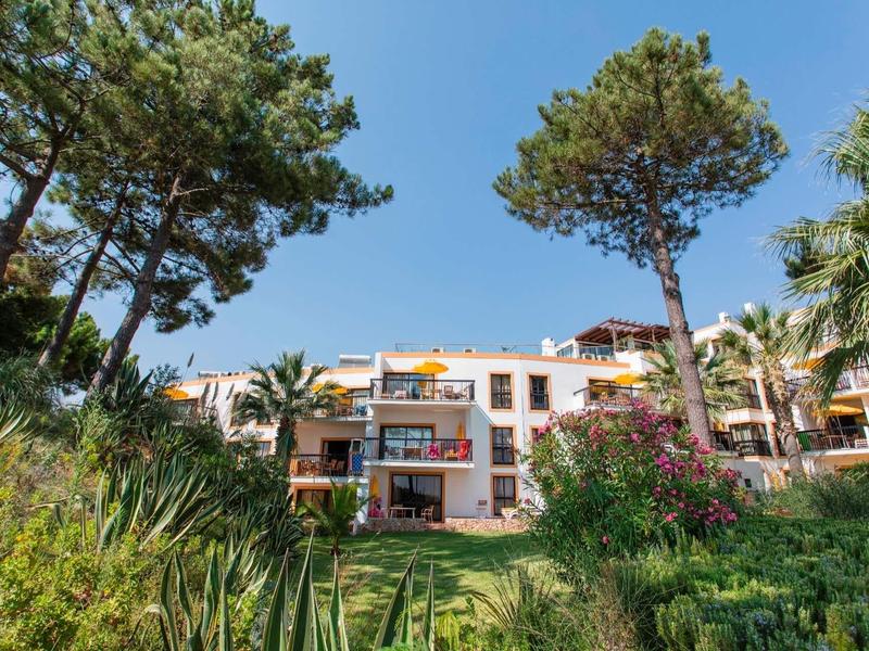 Mehrstöckiges Wohnhaus mit Balkonen, umgeben von Palmen und Kiefern unter blauem Himmel.