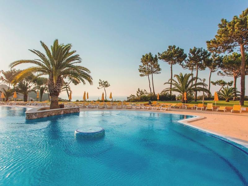 Großer blauer Pool mit Palmen davor und klarem Himmel im Hintergrund, sonnige Atmosphäre.
