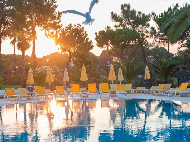 Outdoor-Pool mit blauen Liegestühlen, gelben Sonnenschirmen, Bäumen und Vogel im Sonnenuntergang.