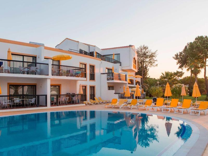 Moderner Hotelbau mit weißen Wänden, großen Fenstern und gelben Sonnenschirmen am klaren Pool.