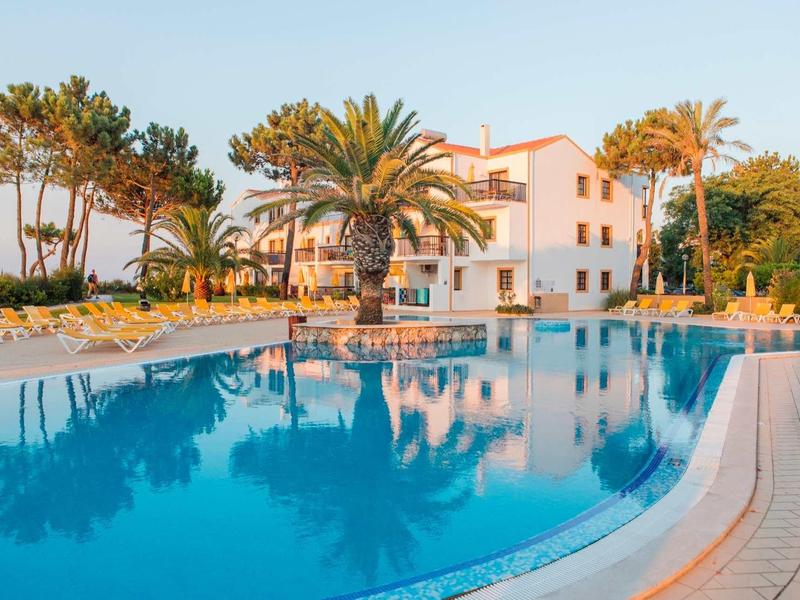 Großer Pool mit klarem Wasser, Palmen und mehrstöckigem weißen Gebäude im Hintergrund bei Sonnenuntergang.