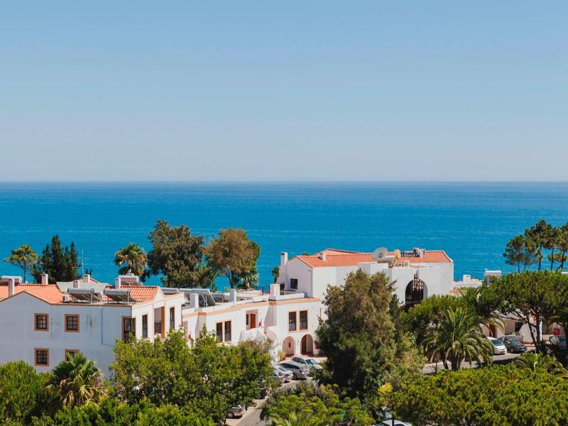Weiße Häuser mit roten Dächern, umgeben von grünen Bäumen, Meer und blauem Himmel im Hintergrund.