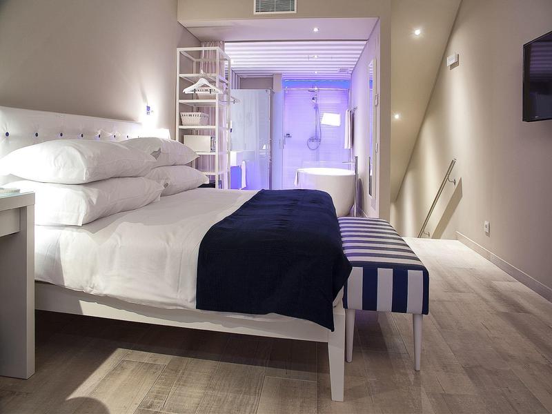 Modernes Schlafzimmer mit weißem Bett, dunkelblauer Decke, gestreifter Bank und beleuchtetem Badezimmer.