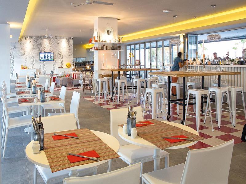 Moderner Gastraum mit weißen Möbeln, Holztischen, roten Servietten und großen Fenstern.