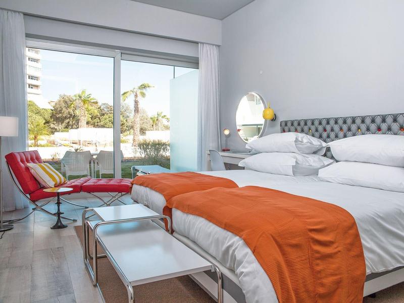 Helles Schlafzimmer mit großem Bett, orangener Decke, weißem Nachttisch und Gartenausblick durch Glasfenster.